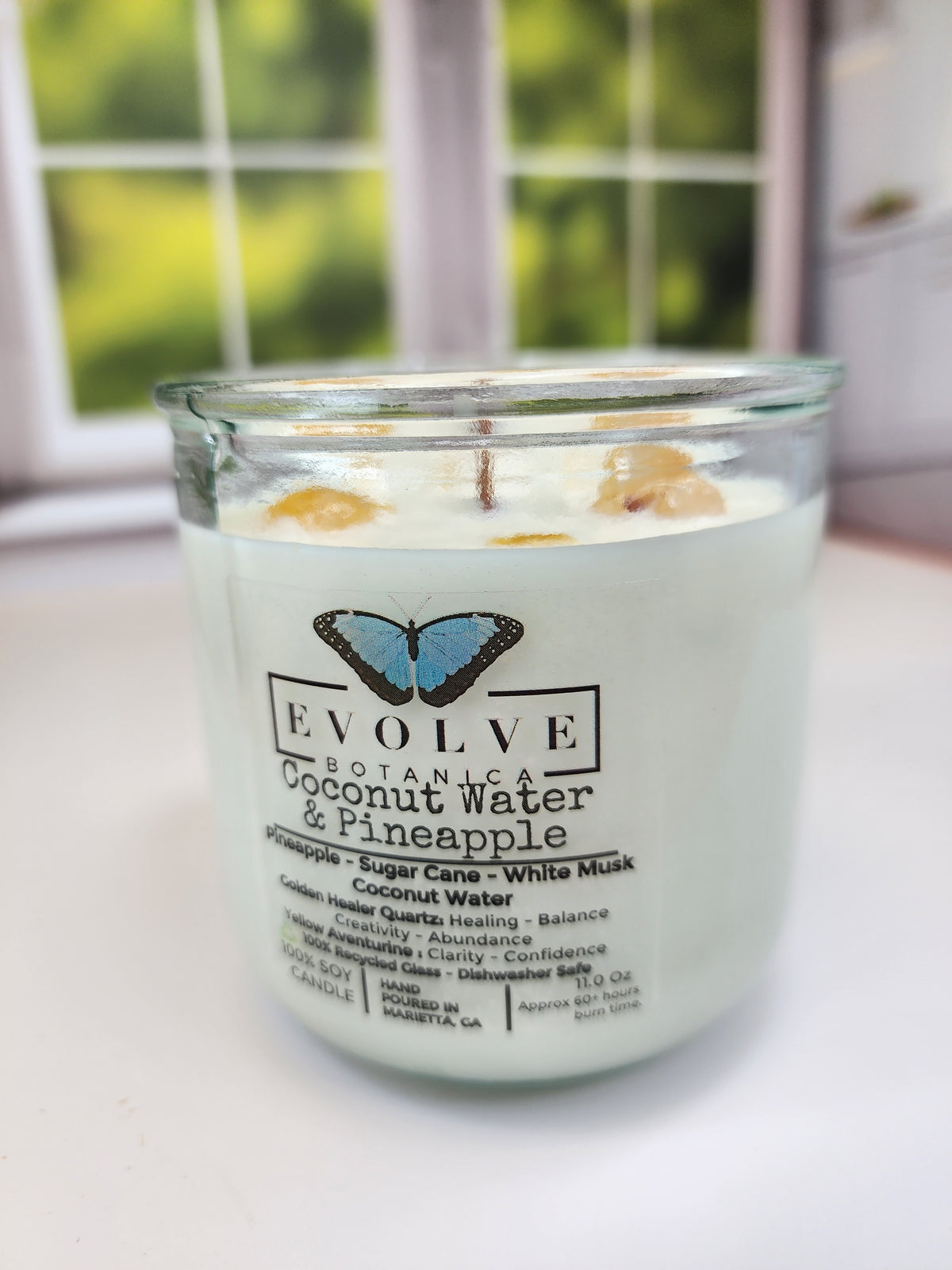 Coconut Water & Pineapple Rocks candle on a windowsill in daylight — tropical wood wick soy candle topped with golden healer quartz and yellow aventurine chips