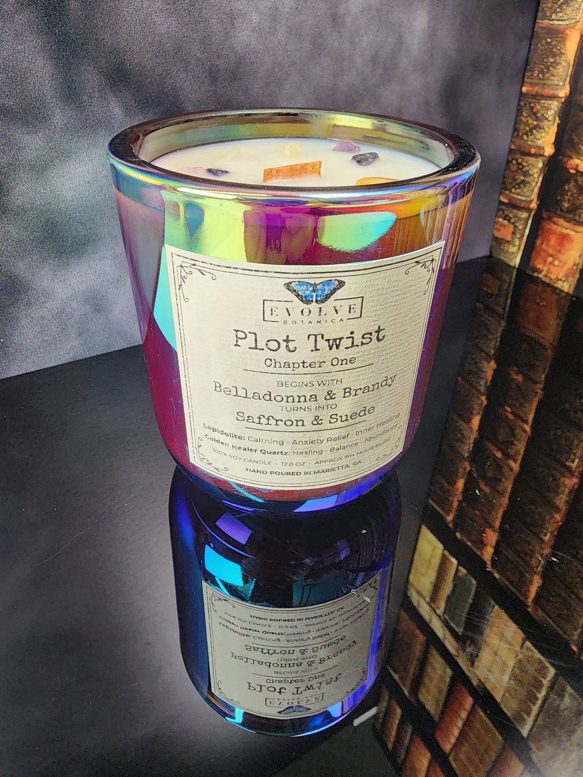 Plot Twist Chapter One 12 oz iridescent glass candle on a reflective surface, Belladonna & Brandy to Saffron & Suede layered wood wick gemstone candle