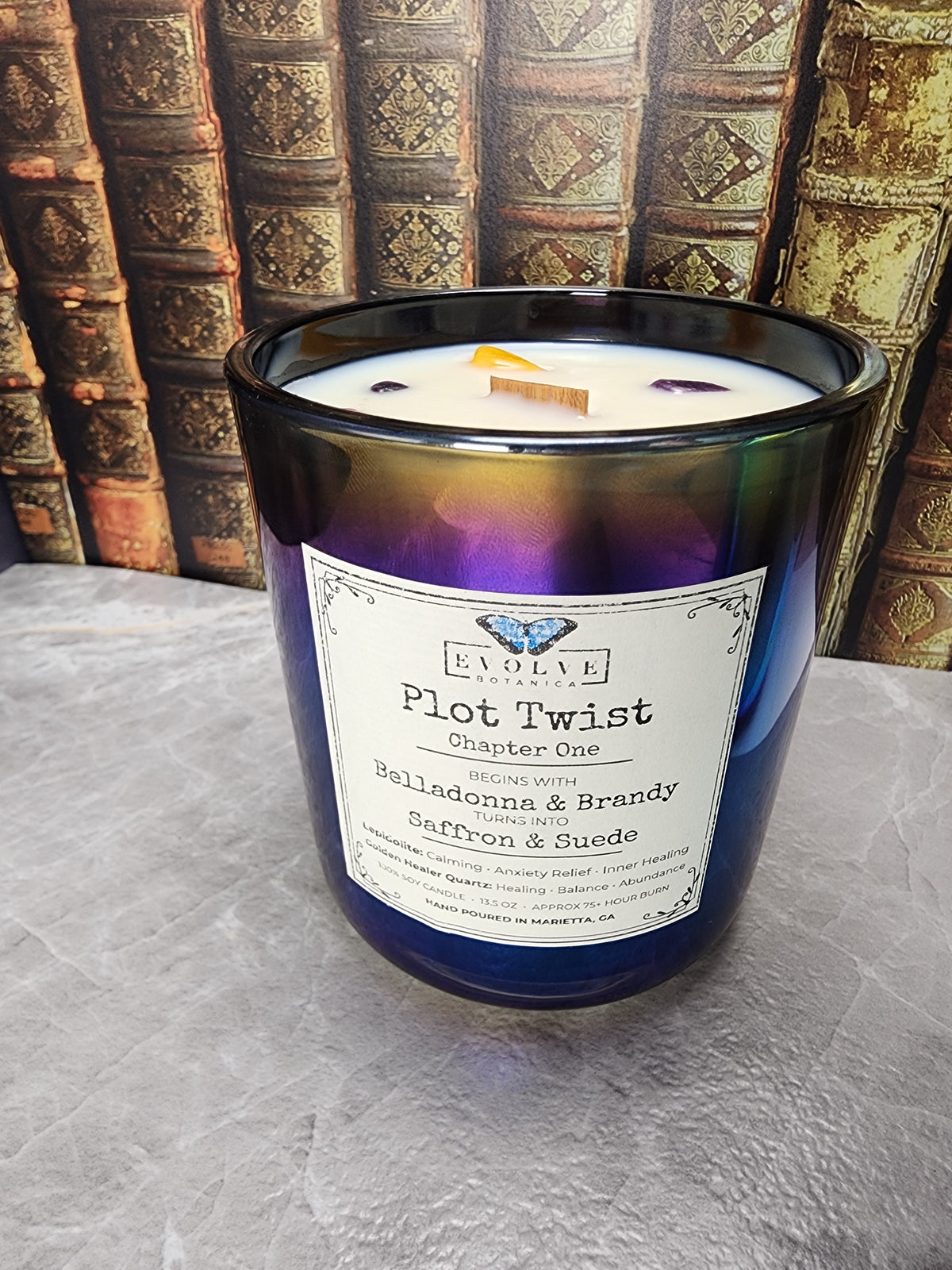 Plot Twist Chapter One 13.5 oz layered soy candle in iridescent black glass with wood wick and Lepidolite and Golden Healer Quartz, against antique leather-bound books