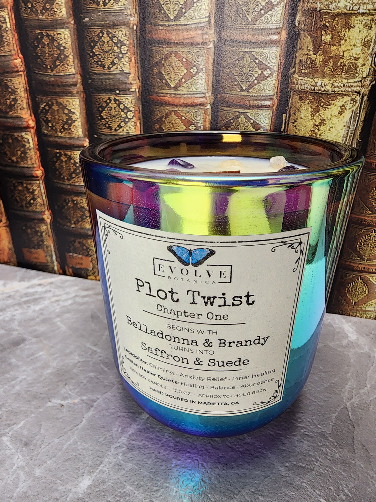 Plot Twist Chapter One 12 oz iridescent glass gemstone candle with filigree label in front of a wall of antique books, bookish limited-edition candle