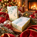 Two Silver Spruce shea butter soap bars with green and cream swirls among frosted pine, red velvet ribbon and ornaments by a fireplace